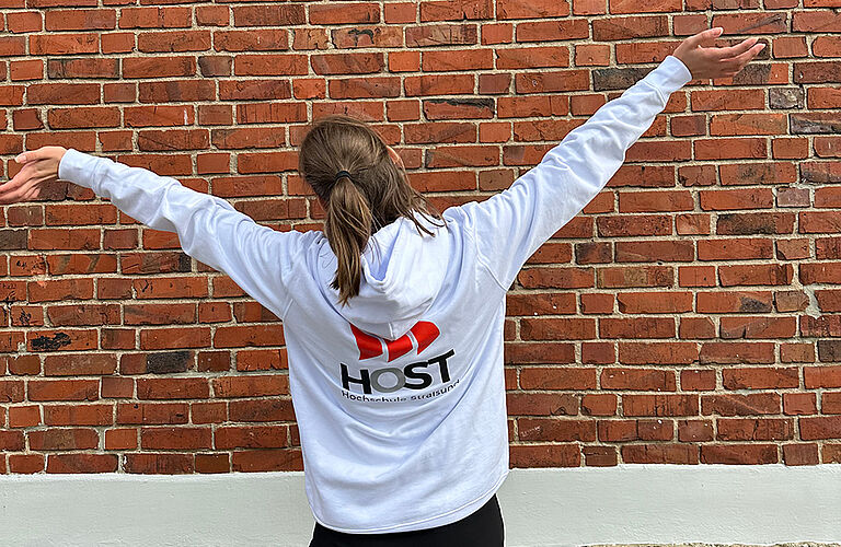 dunkelhaarige Frau von hinten fotografiert, die einen weißen Hoodie mit HOST-Logo trägt und vor einer Backsteinbauer steht