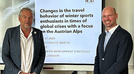 Prof. Jan-Pierre Klage und Martin Breitkreutz stehen in schwarzen Anzügen vor einem Beamerbild mit dem Titel seiner Abschlussarbeit