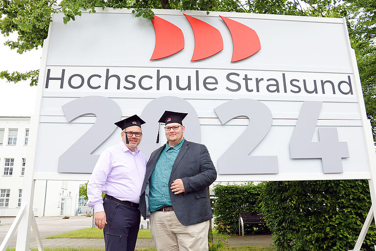 Zwei Männer mit Absolventenkappen stehen vor einem Hochschul-Schild mit einer großen 2024 darauf. 