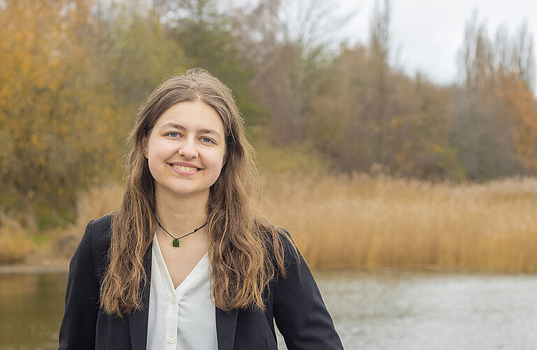 Portraitaufnahme von Julia Koch an einem See mit Bäumen im Hintergrund