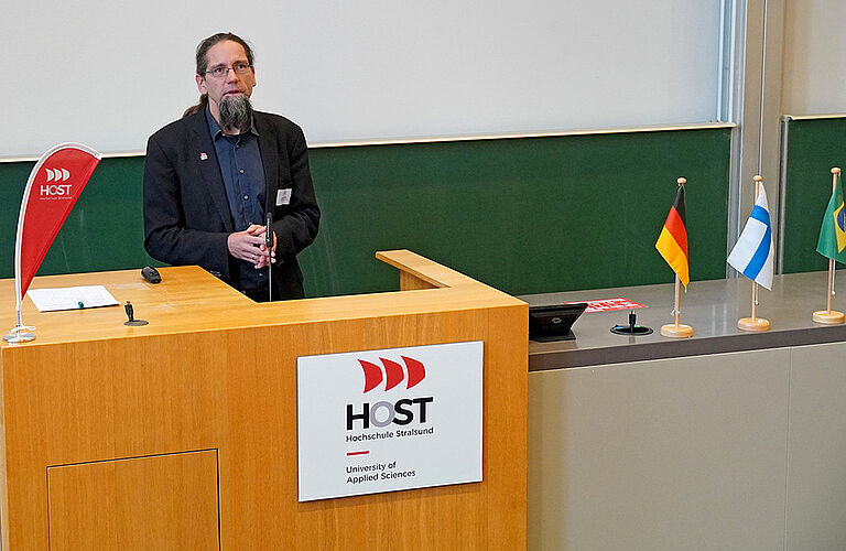 Prof. Johannes Gulden, in schwarzen Anzug, steht an einem Pult mit dem Logo Hochschule Stralsund, vor einer grünen Tafel.