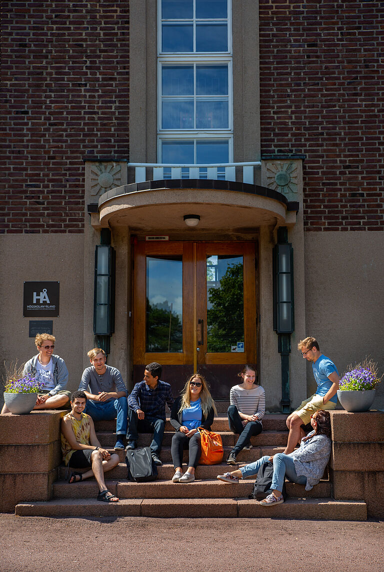 eine Gruppe junger Menschen sitzt vor einem Gebäude, vermutlich einer Fakultät, auf den Treppenstufen.