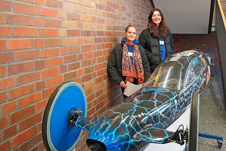 zwei junge Frauen stehen hinter einem sehr schmalen, kleinen Rennwagen mit blauen Blitzen darauf.